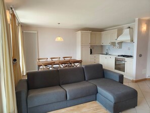 Ein modernes Wohnzimmer mit einem grauen Ecksofa, einem Esstisch mit Stühlen, einer Küche mit weißen Schränken, einem Einbauofen und einer Pendelleuchte.