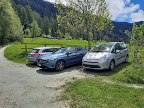 Un sendero pavimentado con coches aparcados, una cerca de madera y montañas al fondo.