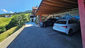 Un área de estacionamiento con un coche blanco y una camioneta SUV negra, un edificio con techo inclinado y vistas de un paisaje verde con casas.