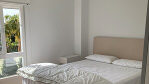 Ein Schlafzimmer mit einem hölzernen Bett, weißer Bettwäsche, einem Nachttisch und einem Fenster mit Blick ins Grüne.