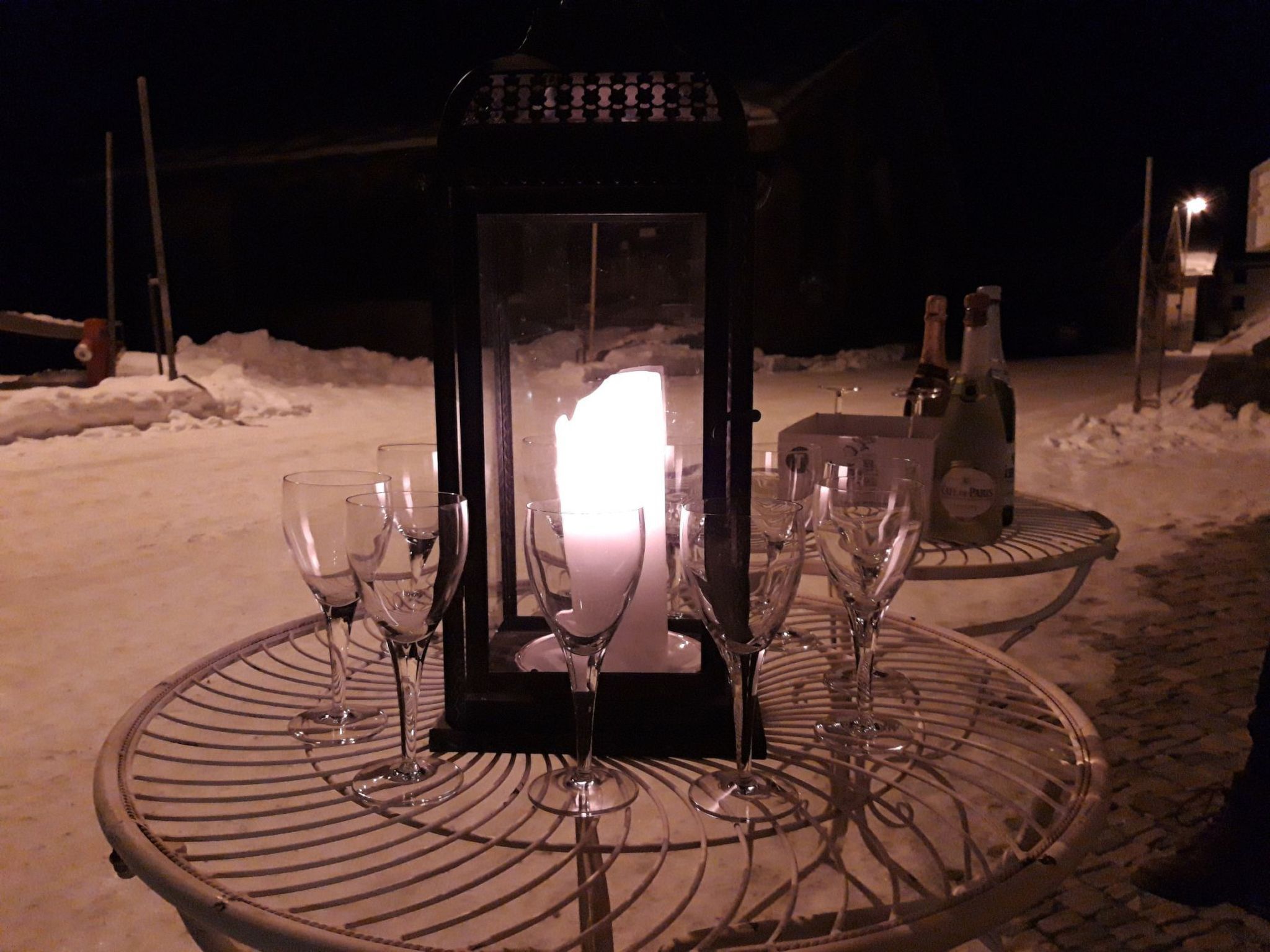 Een besneeuwde omgeving buiten, met een tafel gedekt voor een maaltijd, inclusief een kaars, wijnglazen en een fles.