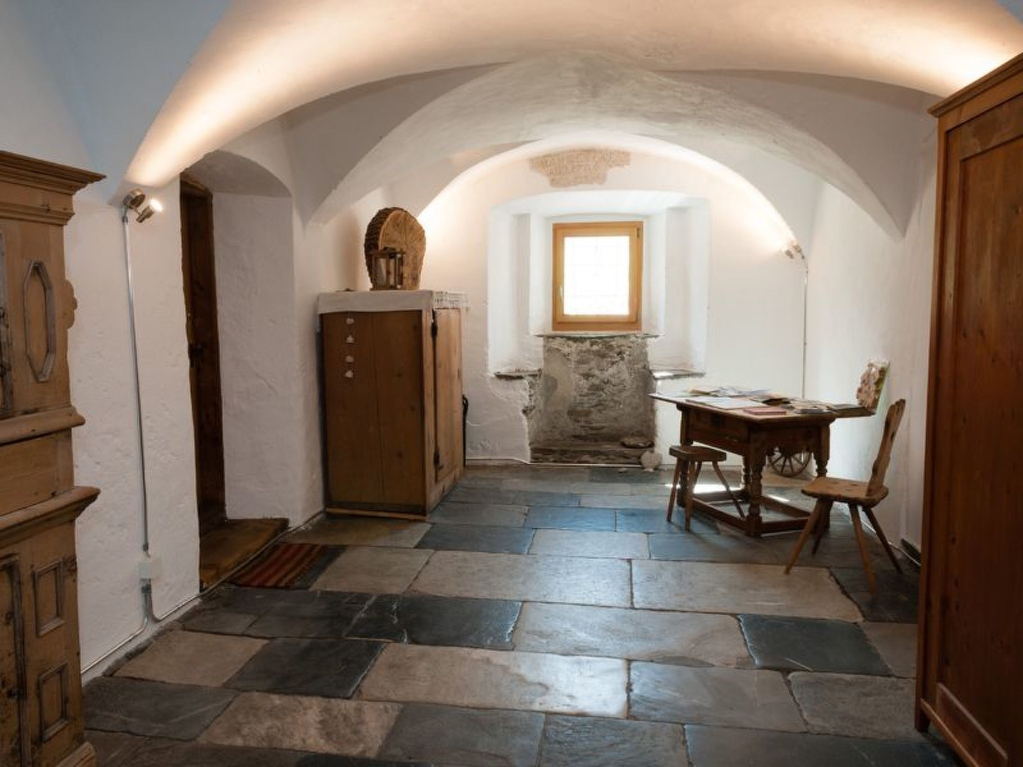 A narrow hallway with stone flooring, a wooden cabinet, a table with a chair, and a window with natural light.