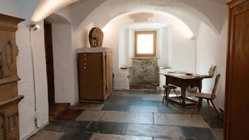 A narrow hallway with stone flooring, a wooden cabinet, a table with a chair, and a window with natural light.