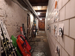 Un couloir étroit où sont appuyés contre le mur des paires de skis et des planches de snowboard, une porte en bois au fond, et un mur recouvert de carreaux orné d’œuvres d’art.