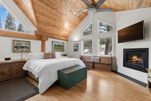 A bedroom with a bed, a bench, a fireplace, and large windows. - Ski-in/Ski-out Cabin w/ Sauna and Hot Tub (Whitefish)