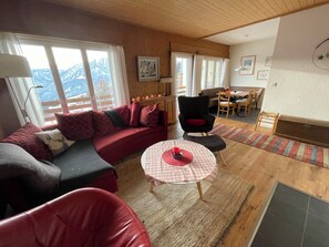 A cozy living area with a sofa, a red chair, a small table with a checkered tablecloth, and a view of the outdoors through large windows. - Wohnung Ohlenroth-Kalberer, (Sarn-Lescha). by Interhome (Sarn)