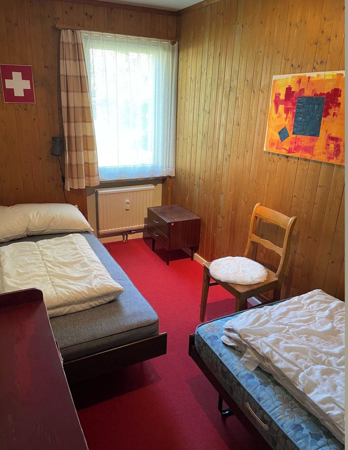 Een kamer met twee bedden, een stoel, een klein tafeltje, een radiator en een raam met gordijnen.