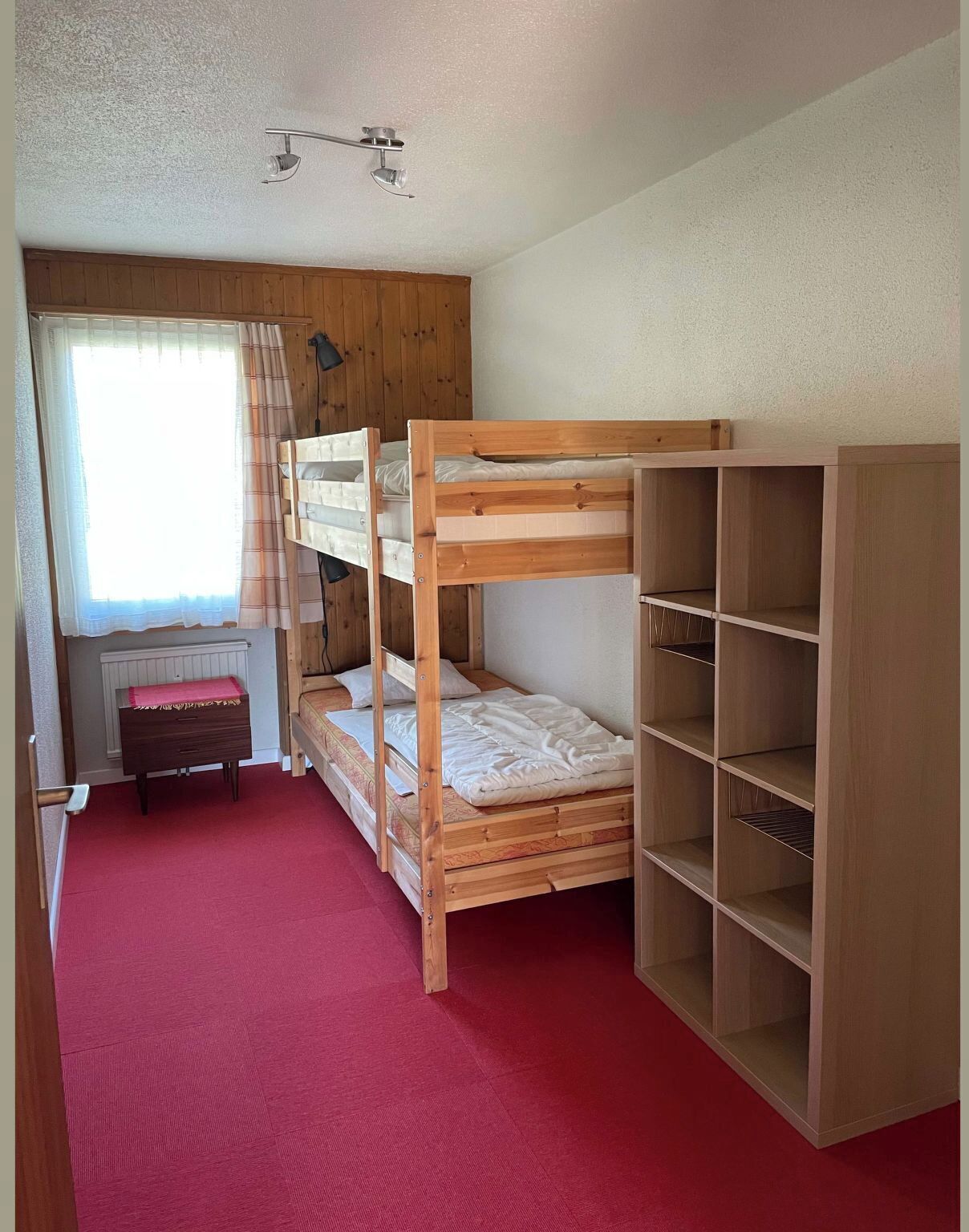 A bunk bed room with a window, radiator, and a wooden wardrobe.