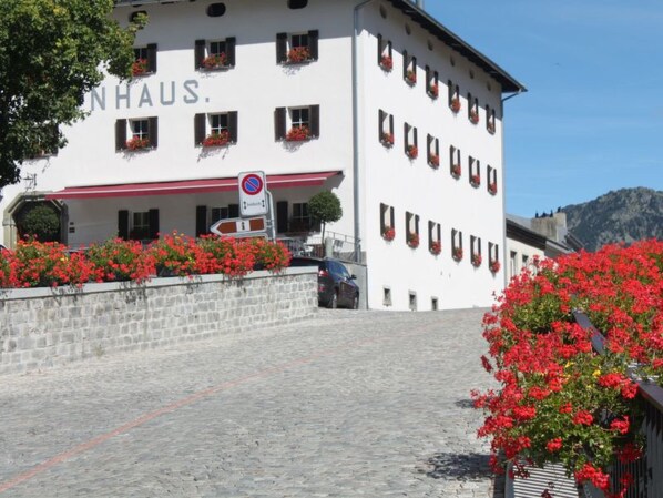 Voici un hôtel dont l’enseigne indique « NHAUS ». Il présente une façade en pierre, de nombreuses fenêtres ornées de jardinières et une entrée dallée.