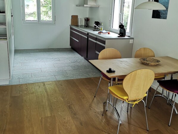 A modern kitchen with a dining area, featuring a large window, a coffee machine, and a wooden dining table with chairs. - Apartment Beeli, (Splügen), by Interhome (Splügen)