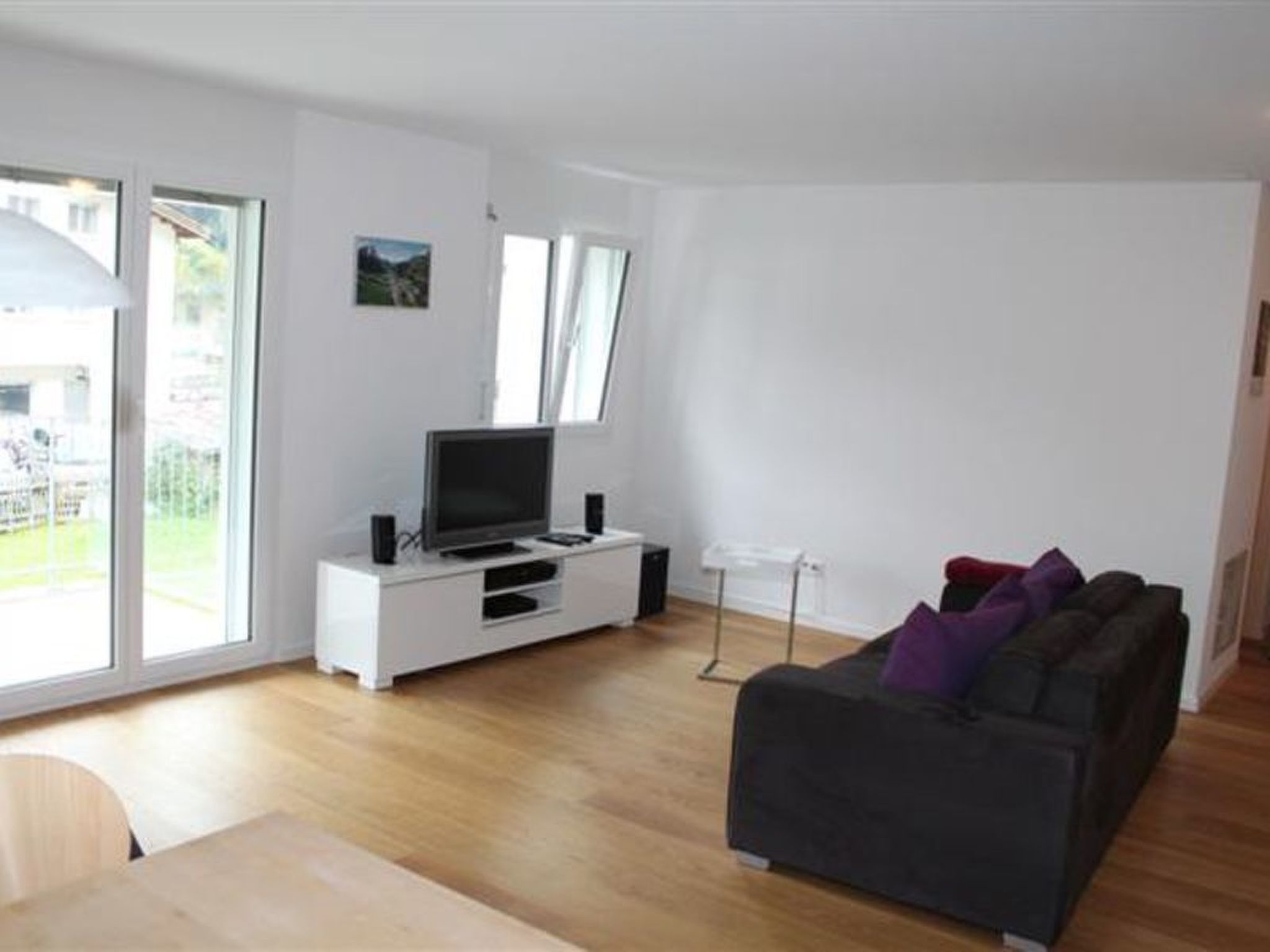 A living room with a TV on a white entertainment unit, a dark sofa with cushions, and a dining table.