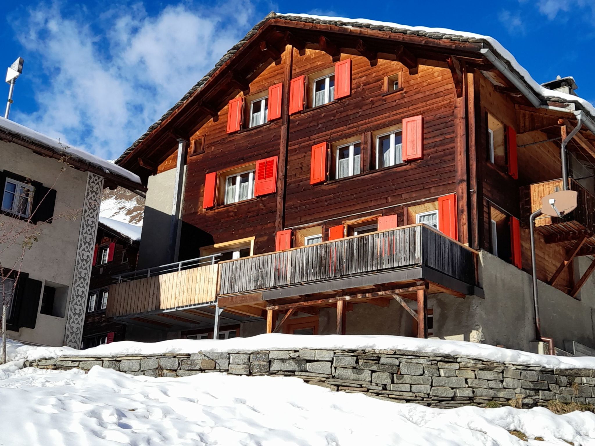 A multi-story wooden building with balconies, a stone wall in front, and a clear blue sky.