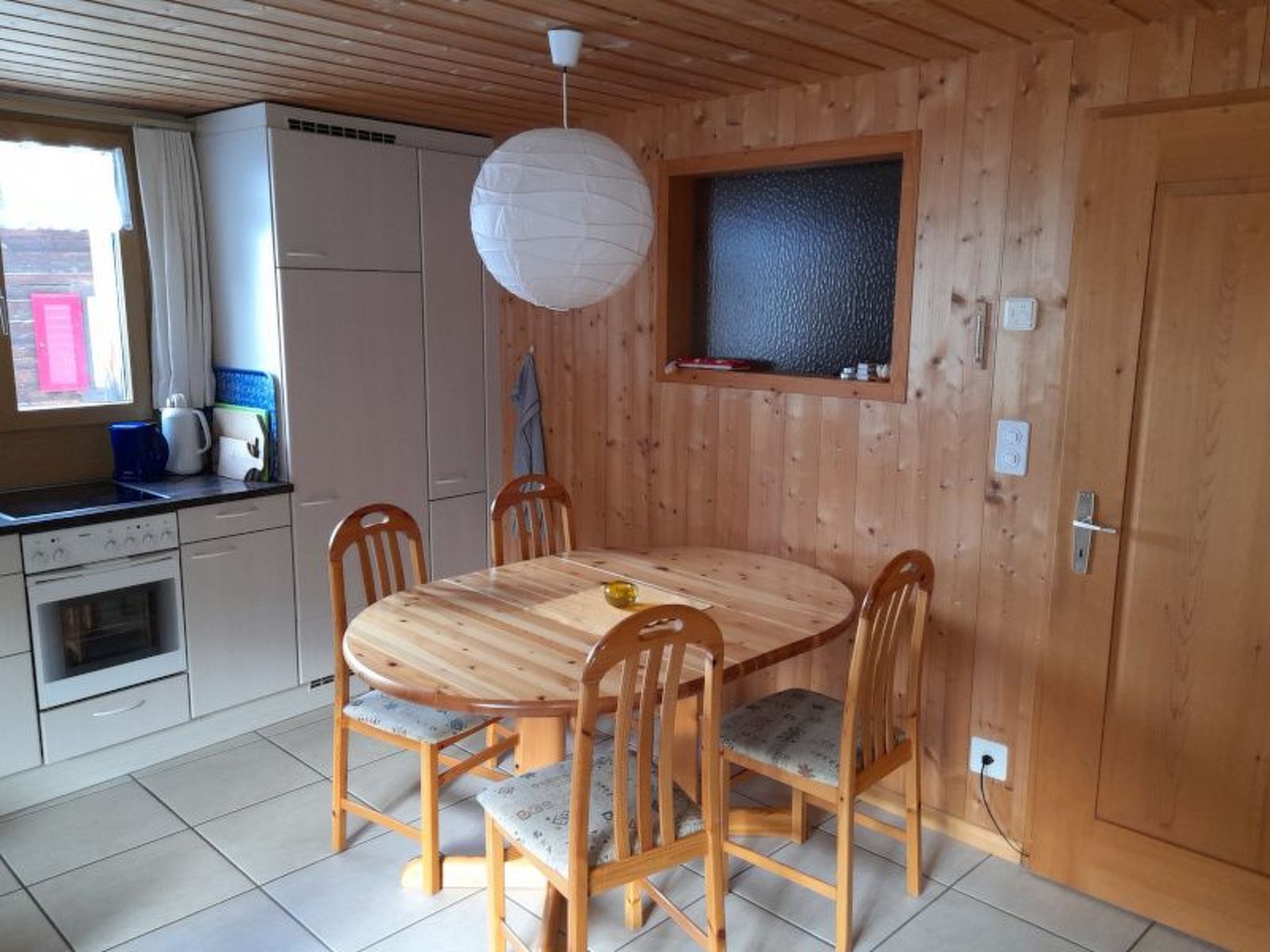 A wooden interior with a dining table, chairs, a hanging light fixture, and a kitchen area.