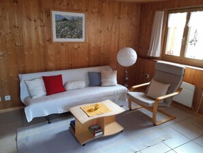 Ein Zimmer mit Holzwänden, einem weißen Sofa, einem Holzstuhl, einem kleinen Tisch mit Lampe und einem Fenster mit Vorhängen.