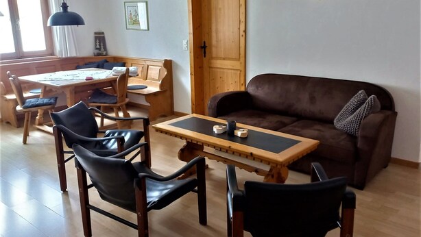 Un salón con un sofá de cuero marrón, una mesa de comedor de madera y sillas, una puerta de madera y adornos de asta en la pared.