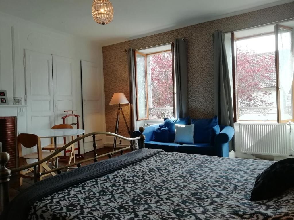 Una habitación con cama, sofá azul, mesa de comedor con sillas, lámpara y ventanas grandes con cortinas.