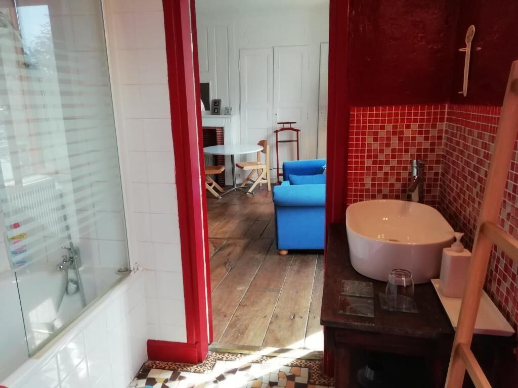 Un baño con bañera, lavamanos y ducha. En la habitación contigua hay una silla azul y una mesa.