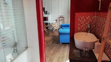 Un baño con bañera, lavamanos y ducha. En la habitación contigua hay una silla azul y una mesa.