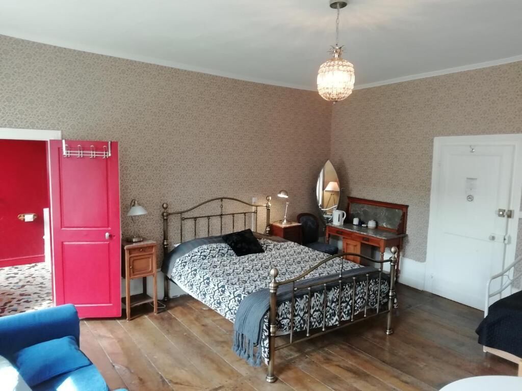 Un dormitorio con cama, mesitas de noche, lámpara, espejo, puerta roja, silla azul y candelabro.