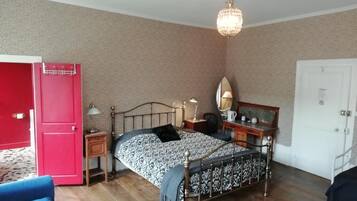 Un dormitorio con cama, mesitas de noche, lámpara, espejo, puerta roja, silla azul y candelabro.