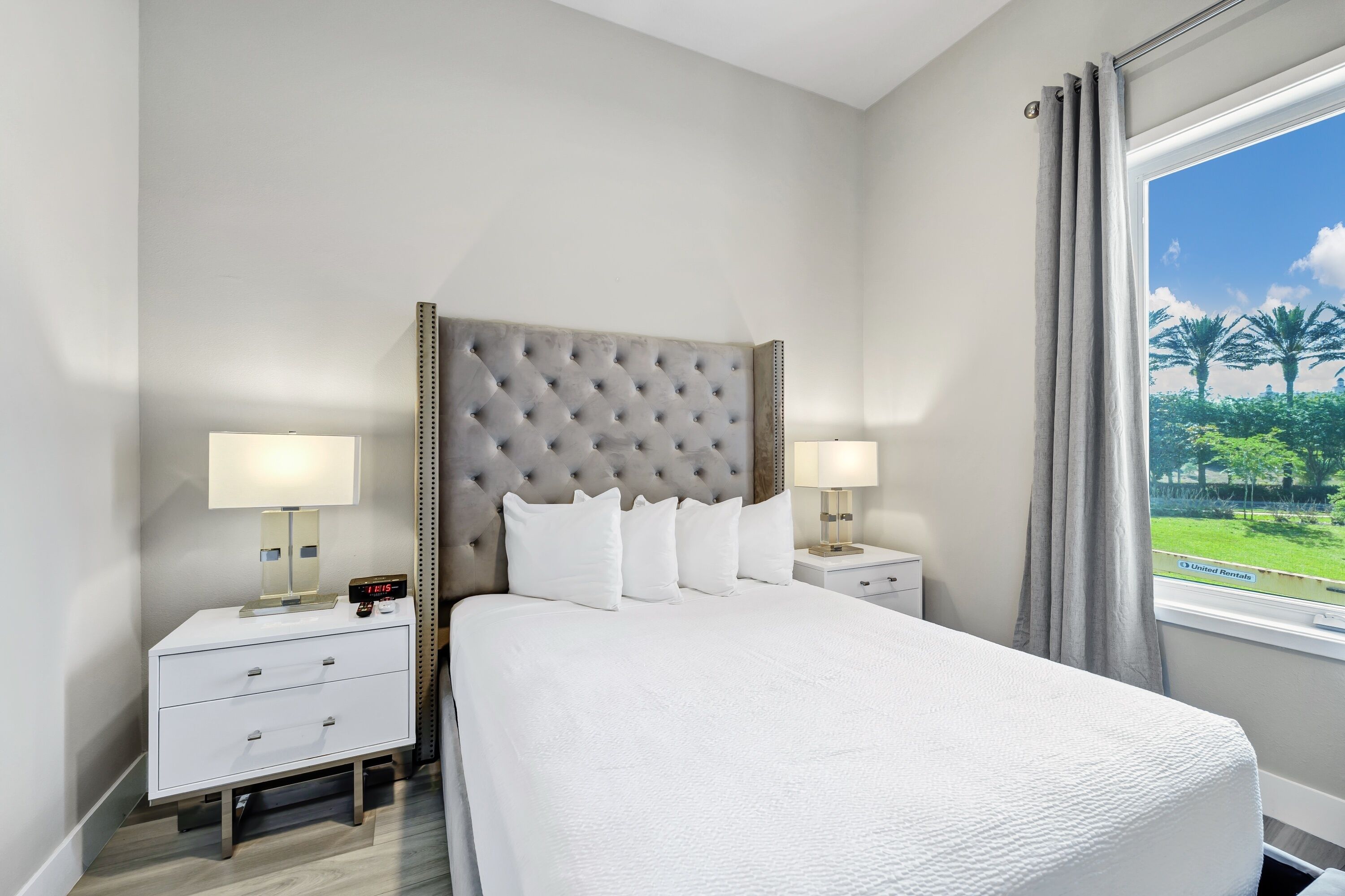 A hotel room with a large bed, two bedside tables, a nightstand, a window with a view of palm trees, and a grey upholstered headboard.