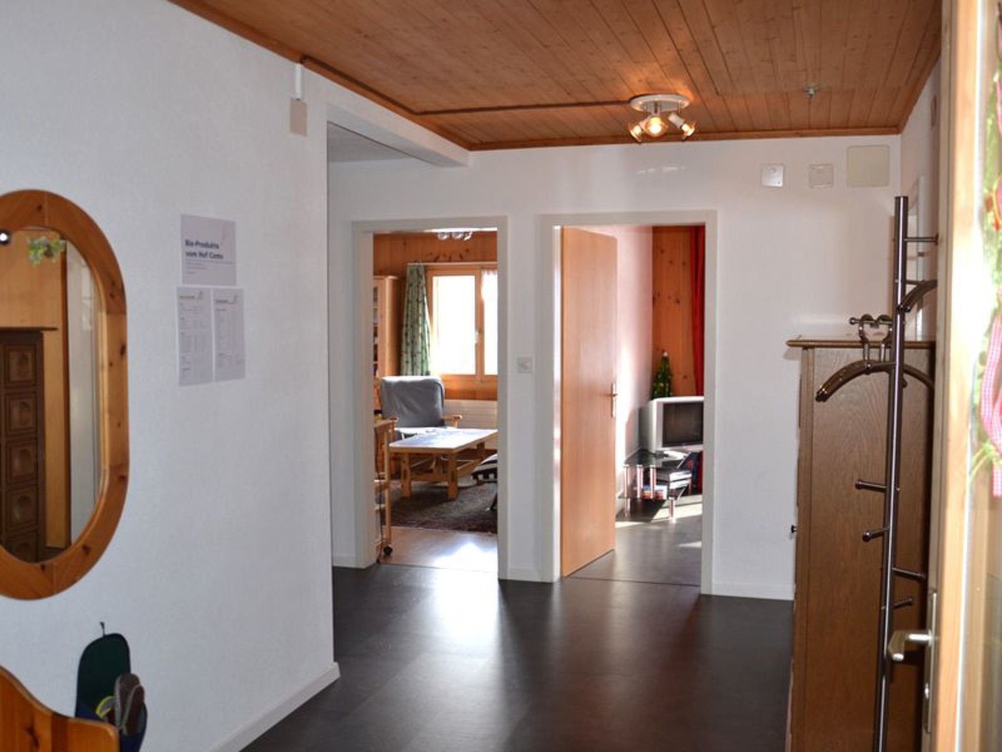 Ein Korridor mit Holzboden, einem Spiegel und einem Kleiderständer. Eine Tür führt zu einem Zimmer mit einem Bett und einem Fernseher.