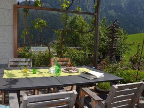 An outdoor dining area with a table set for two, surrounded by wooden chairs, a glass of water, a green bottle, and a book.