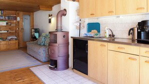 A cozy cabin interior with a wood-burning stove, wooden cabinets, a kitchen area, and a living space with a sofa and television. - Haus Aebli, (Hinterrhein), by Interhome (Hinterrhein)