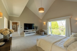 Ein Schlafzimmer mit einem großen Bett, einem Fernseher, einer weißen Kommode, einem Blumenstrauß und Blick auf einen Garten durch ein Fenster.
