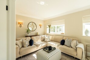 Ein Wohnzimmer mit einem beigefarbenen Sofa, einem runden Spiegel, Wandleuchten, einem Fenster mit einer Pflanze und einem gestreiften Hocker.