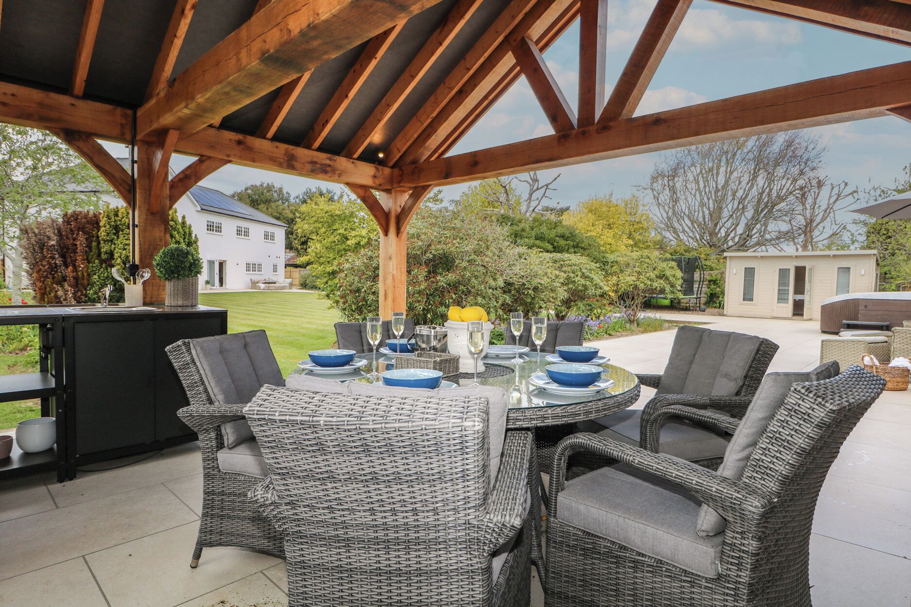 Um pátio externo com mesa posta para refeições, cadeiras de vime e vista para uma casa e um jardim.