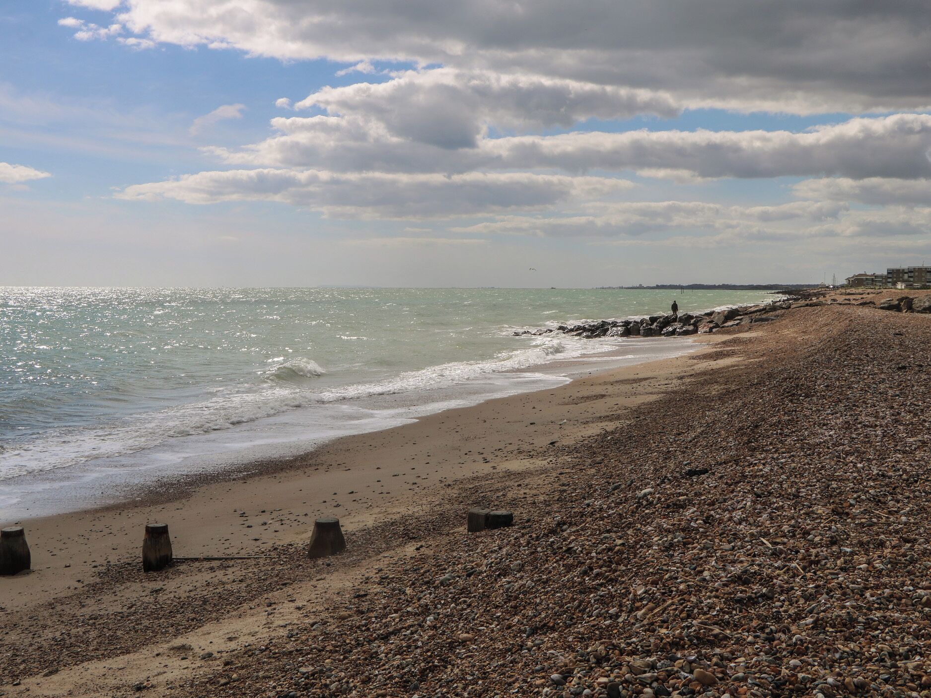 A pebbled beach with a rocky shoreline, a clear sky, and distant buildings.