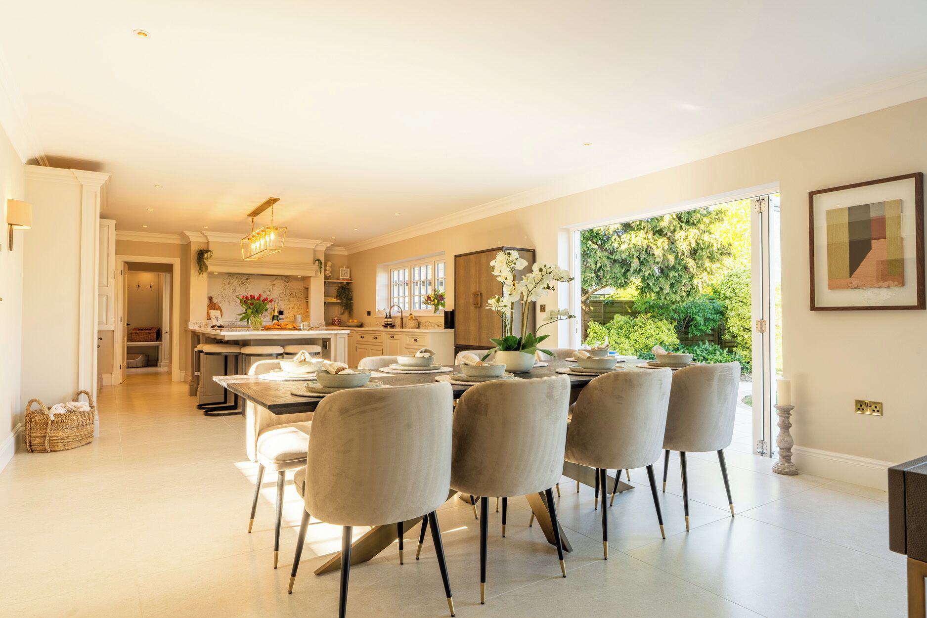 A spacious dining area with a large table set for six, a kitchen in the background, and a view of greenery outside.