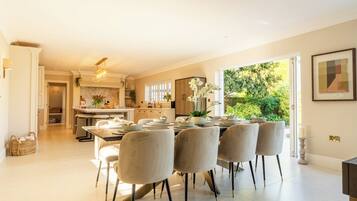 A spacious dining area with a large table set for six, a kitchen in the background, and a view of greenery outside.