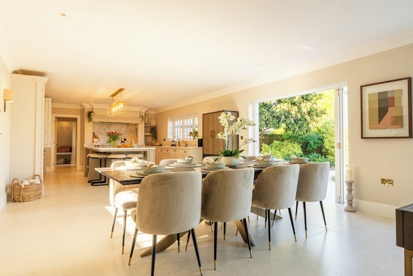 A spacious dining area with a large table set for six, a kitchen in the background, and a view of greenery outside.