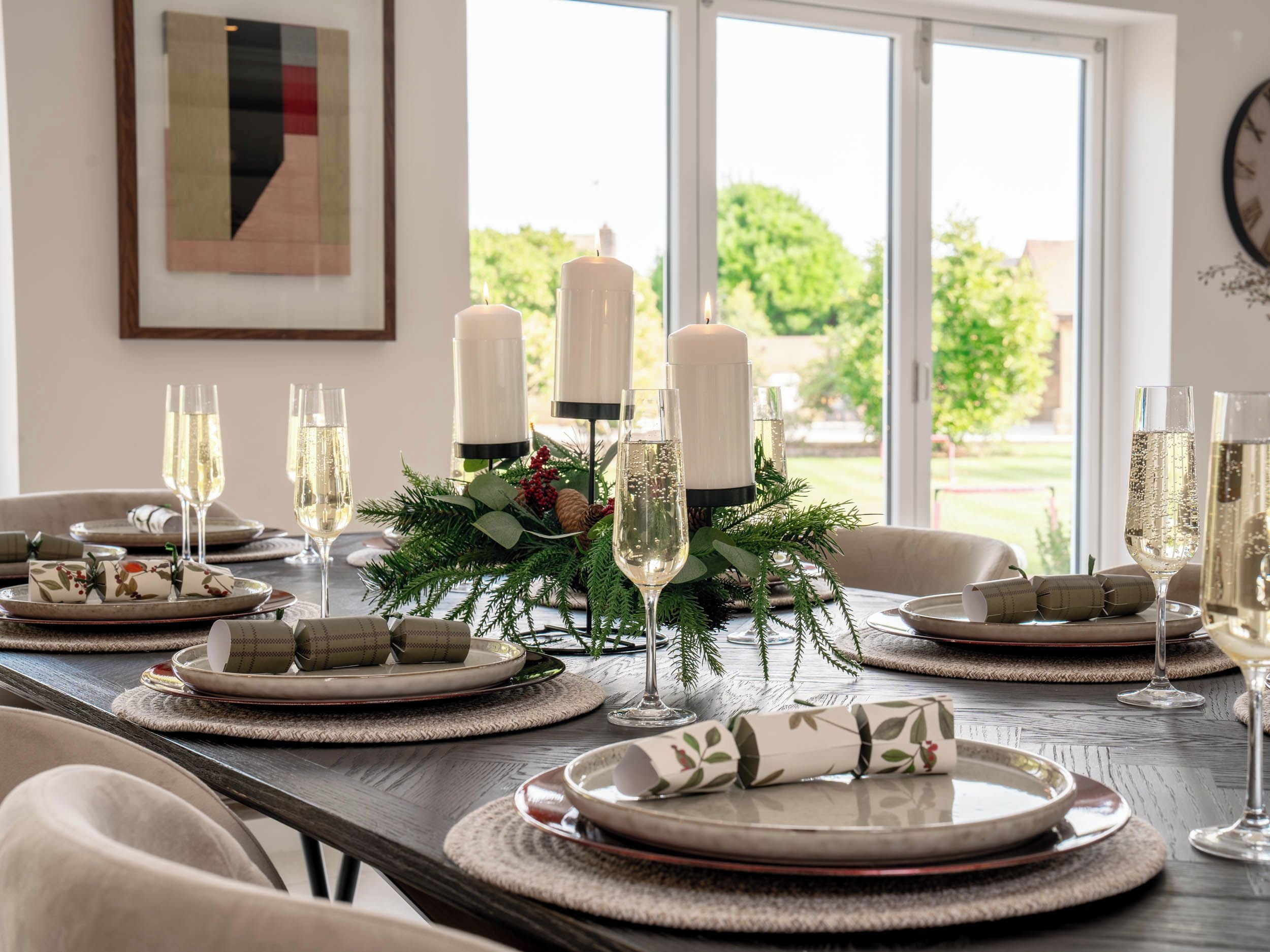 Mesa de comedor dispuesta con platos, vasos y velas, con vistas a la vegetación exterior.