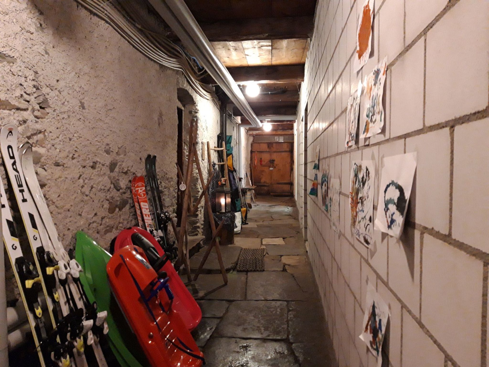 Un couloir étroit où sont appuyés contre le mur des paires de skis et des planches de snowboard, une porte en bois au fond, et un mur recouvert de carreaux orné d’œuvres d’art.