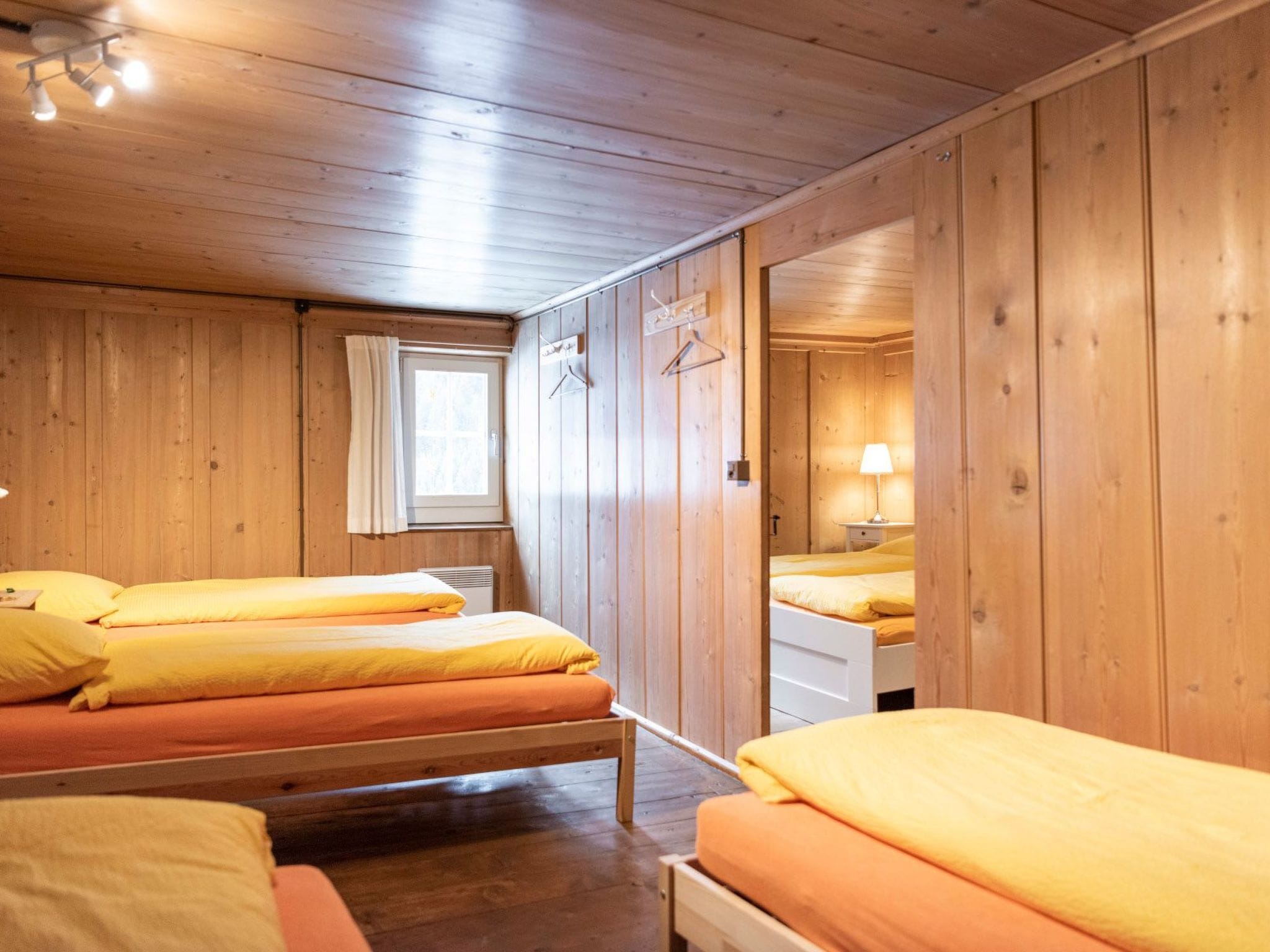 Una habitación con paredes y suelos de madera, dos camas con ropa de cama amarilla, una ventana con cortinas blancas y una lámpara de noche.