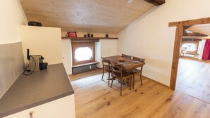 A room with a wooden ceiling, a dining table with chairs, a window with a view, and a small kitchen area. - Wohnung von Schorsch, (Splügen), by Interhome (Splügen)