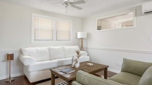 Ein Wohnzimmer mit einem weißen Sofa, einem grünen Sessel, einem Holztisch, einer Stehlampe und einem Deckenventilator.