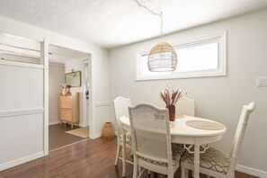 A dining area with a round table, white chairs, a hanging light fixture, and a window with a white frame. - Cozy Coastal Retreat Near Beach & Dining (Englewood)