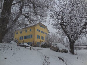A yellow building with multiple windows, surrounded by snow-covered trees and parked cars. - Haus Noains, (Rodels), by Interhome (Sils im Domleschg)