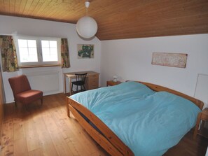 Ein Schlafzimmer mit einem hölzernen Bett, einem Schreibtisch, einem Stuhl, einem Heizkörper und einem Fenster mit Vorhängen.