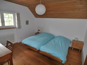 A bedroom with a wooden floor, a bed with a blue bedspread, a nightstand, a desk, and a window with curtains. - Haus Noains, (Rodels), by Interhome (Sils im Domleschg)