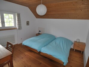 Ein Schlafzimmer mit Holzbalkendecke, einem Bett mit blauer Bettwäsche, einem Nachttisch, einem Stuhl und einem Fenster mit Vorhängen.