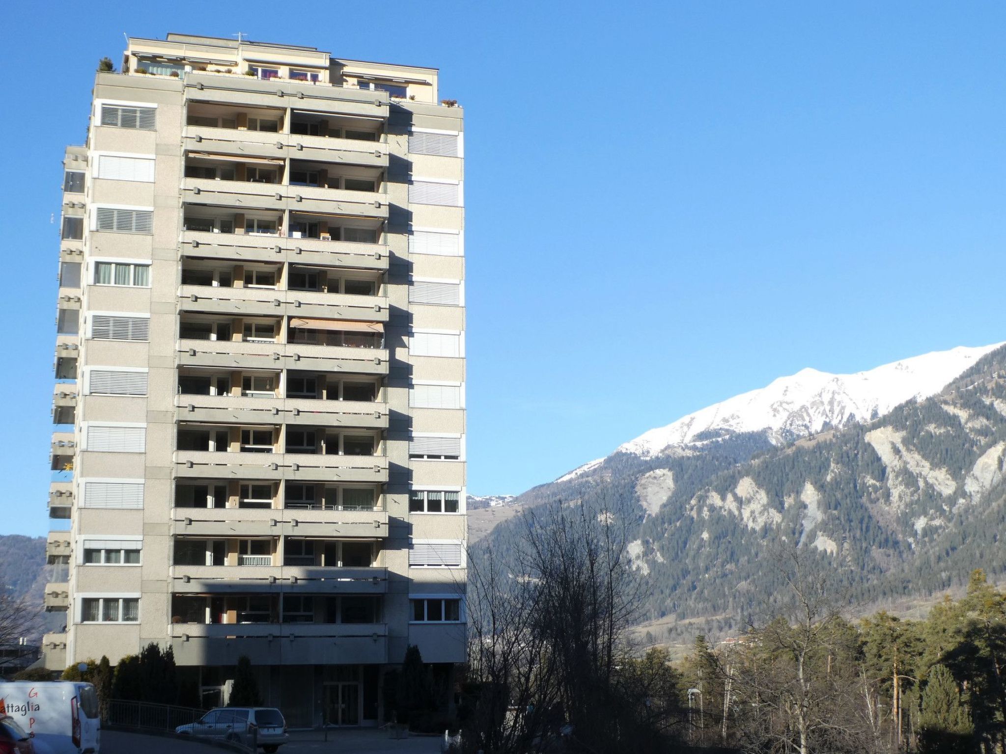 Edifício com várias andares, varandas, área de estacionamento e montanhas ao fundo.