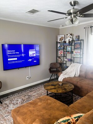 A living room with a flat-screen TV displaying a welcome message, a ceiling fan, a bookshelf with board games, and a brown sofa. - Welcome to The Lone Star Stay! Relaxing 3/2 family/group retreat near downtown! (Kirby)