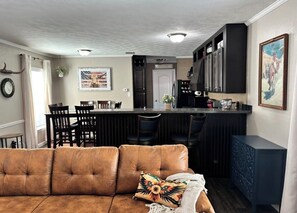 Ein Wohnzimmer mit braunem Sofa, eine Küche mit Bar und ein Essbereich mit Stühlen.