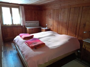 A wooden-paneled bedroom with a bed, bedside tables, a dresser, and a window. - Ferienwohnungen Jörihus A&B, (Medels), by Interhome (Splügen)