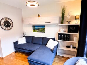 Un salon moderne avec un canapé d’angle, une horloge murale, un tableau, une étagère avec des verres et une lampe.